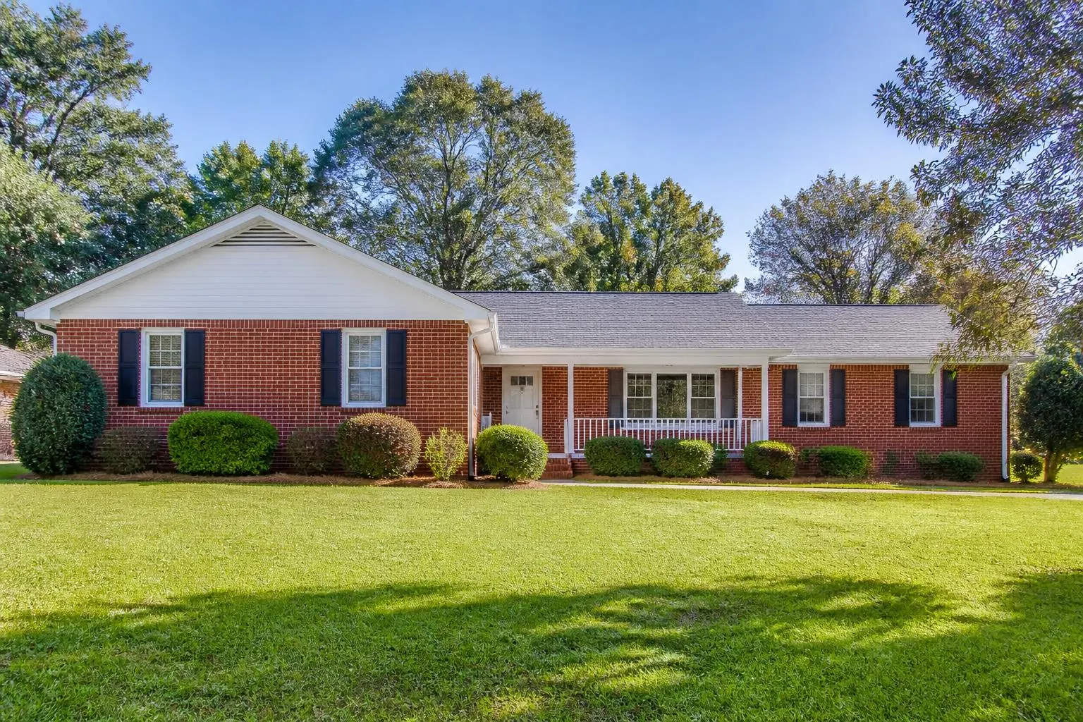 A classic 1960s red brick ranch house with white trim, black shutters and a wide front lawn